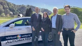Four people stand next to a white automated vehicle. The man on the left wears a suit and tie. Behind him, a woman in glasses and a scarf. The others wear business suits.