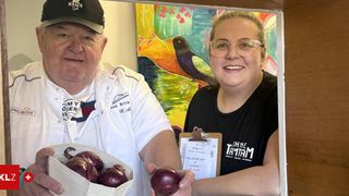 A man and a woman stand with a basket of red onions, smiling and posing for a picture. The man wears a baseball cap and a white shirt, while the woman wears glasses and a black shirt. Behind them is a colorful painting with a bird and leaves.
