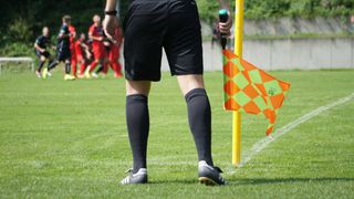 A soccer referee stands on a field holding a whistle and a flag. Players in red uniforms run in the background.