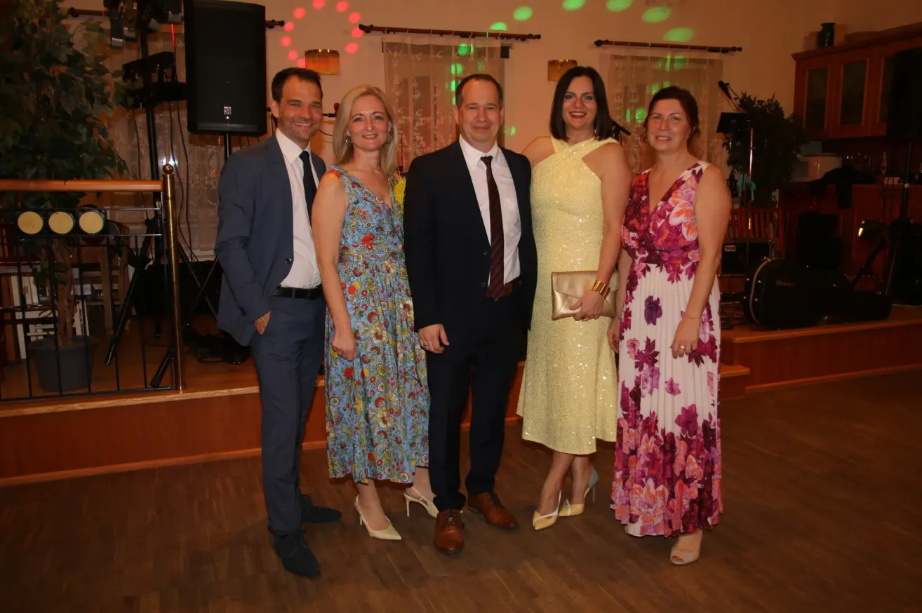 Five individuals, two men and three women, are standing close together on a wooden floor. They are dressed in formal attire and are smiling for a photo. The background features a stage with speakers and lights.