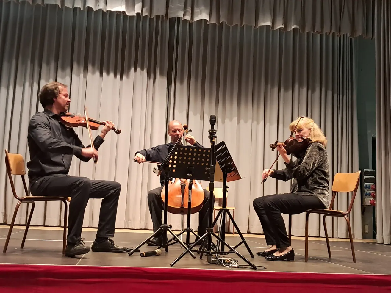 Three musicians on stage, two men and a woman, playing violins and a cello. Behind them, a curtain and a microphone stand.