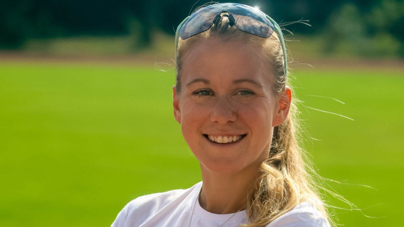 A woman with long blonde hair and sunglasses on her head is smiling in an outdoor area. She is wearing a white shirt and a necklace. The background is blurry.
