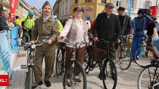 A group of people dressed in vintage clothes stands with their bicycles on a city street. One wears a military uniform, while others wear hats and glasses.