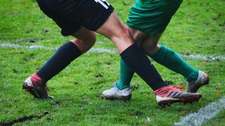 Two soccer players in black and green jerseys are fighting for the ball. The player in black has the number 19 on their shorts.