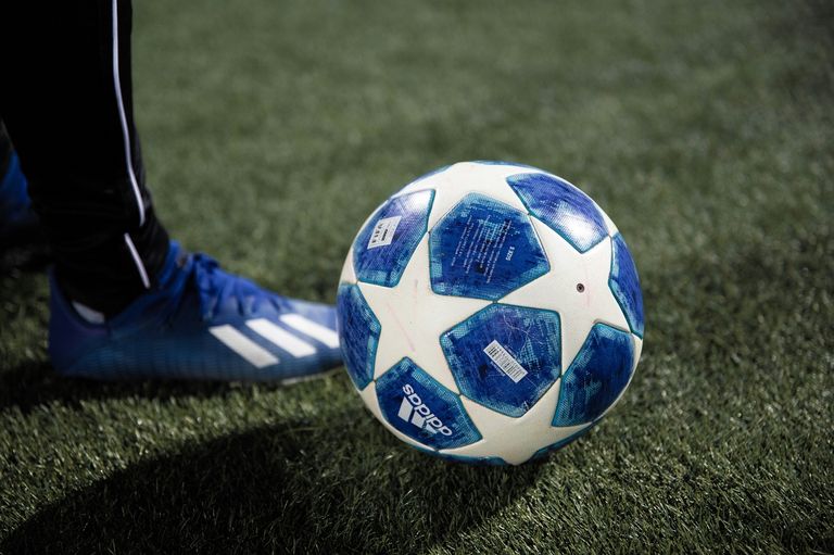 A close-up of a blue and white soccer ball with Adidas branding, partially covered by a blue shoe on a green field.