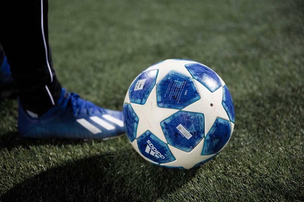 A close-up of a blue and white soccer ball with Adidas branding, partially covered by a blue shoe on a green field.