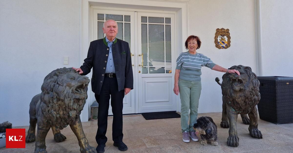 An elderly couple stands with a dog next to two lion statues, posing for a photo in front of a white door with glass panels and a crest on the wall.