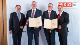 Four men stand posing for a photograph. Two of them hold certificates. Behind them is a backdrop with logos.
