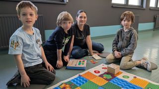 Four children sit on the floor playing a board game, with a woman smiling and a boy focused on the game.