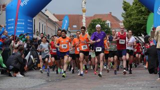 Participants in a marathon run on a city street, wearing numbered bibs. Buildings and greenery form the backdrop.