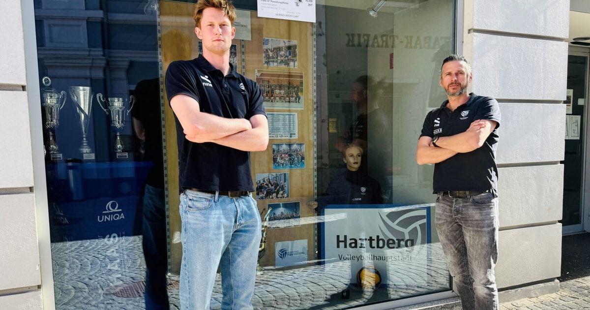 Two men stand in front of a sports shop. One wears a black shirt and jeans, the other a black shirt with a logo. They both have their arms crossed. Behind them, a mannequin and trophies are visible through the shop window.