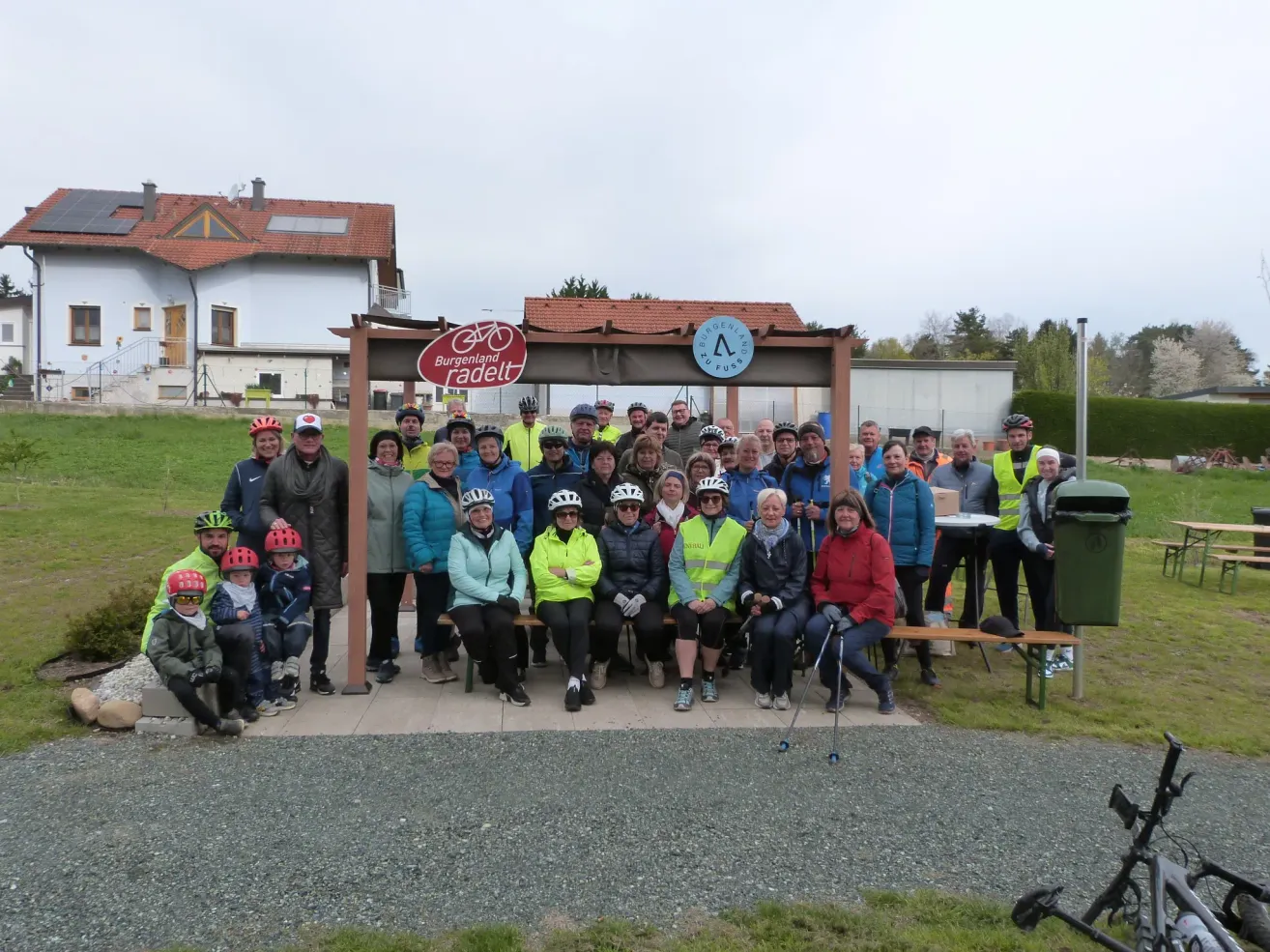 A group of people in cycling gear and helmets gather outside a building with a red sign and bicycle symbol. Behind them is a building with a brown roof.