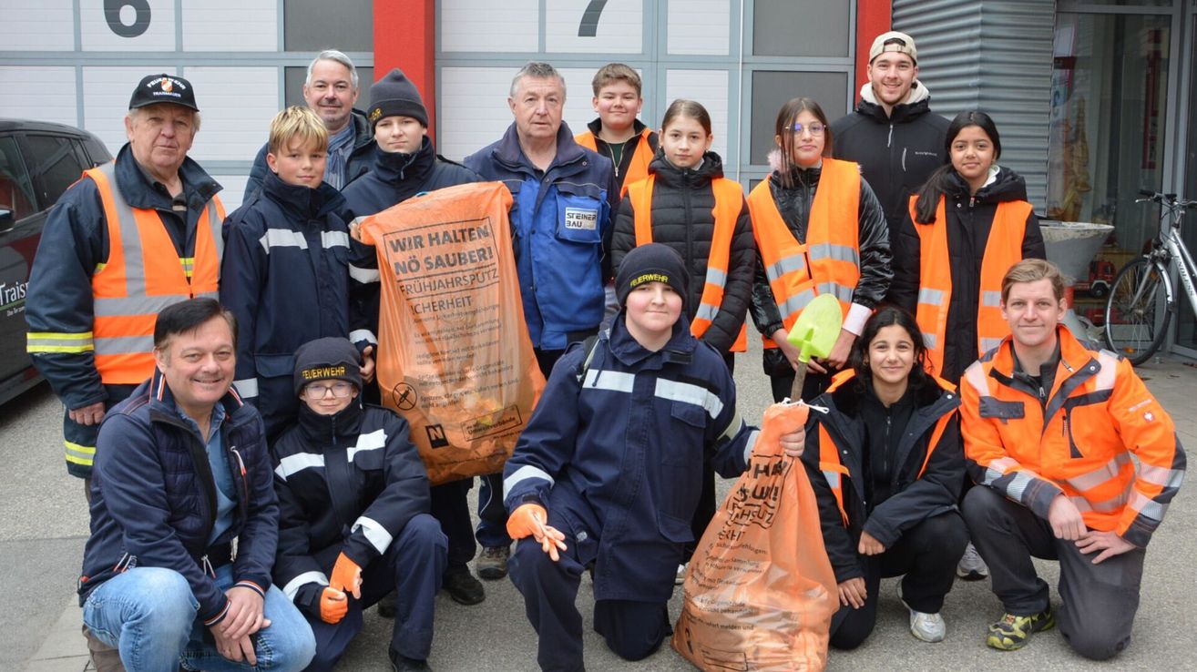 A group of people in reflective vests are posing for a photo, holding orange garbage bags. They are outside a building with red walls.