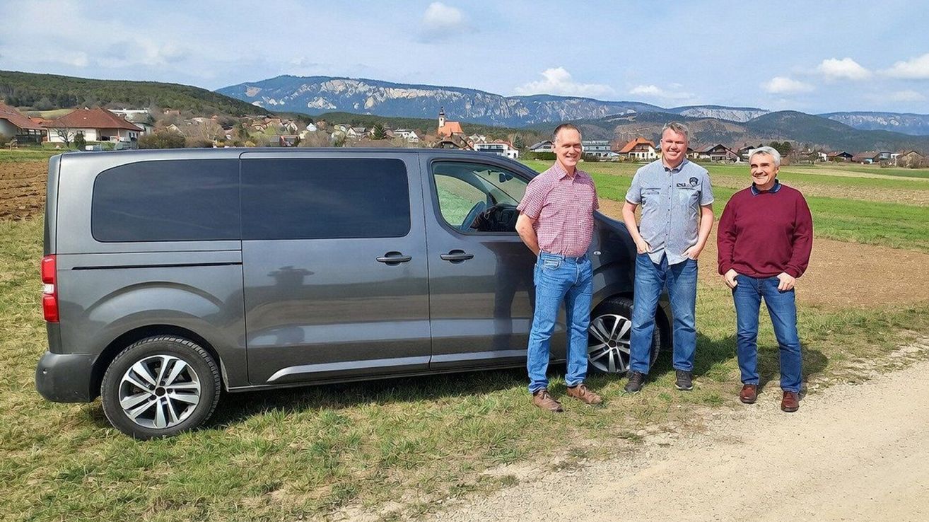 Two men stand in front of a gray van in a rural area, with mountains and a small village in the background.