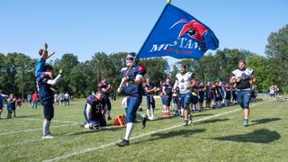 Fußballspieler in Uniform halten eine Flagge während eines Spiels. Ein Spieler kniet auf dem Feld, während andere hinter ihm stehen. Bäume und ein blauer Himmel sind im Hintergrund.