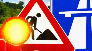 Ein Verkehrsschild warnt vor bevorstehenden Straßenarbeiten mit einer Silhouette eines Arbeiters und einer gelben Leuchte.
