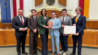 Eine Gruppe von Menschen in formeller Kleidung steht nebeneinander, lächelt und hält Urkunden in der Hand. Ein Mann in einem hellblauen Anzug hält einen Preis. Hinter ihnen steht ein Holzschreibtisch mit einem roten Vorhang.