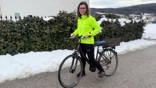 Eine Frau steht neben einem Fahrrad mit Korb, trägt eine leuchtend gelbe Jacke und eine Brille. Hinter ihr steht eine schneebedeckte Hecke und eine Straße.