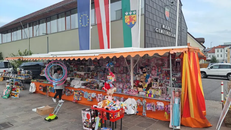 A stall selling toys is located in front of the Market Gemeindeam building with flags of Europe, Austria, and a local coat of arms. A woman is looking at the toys.