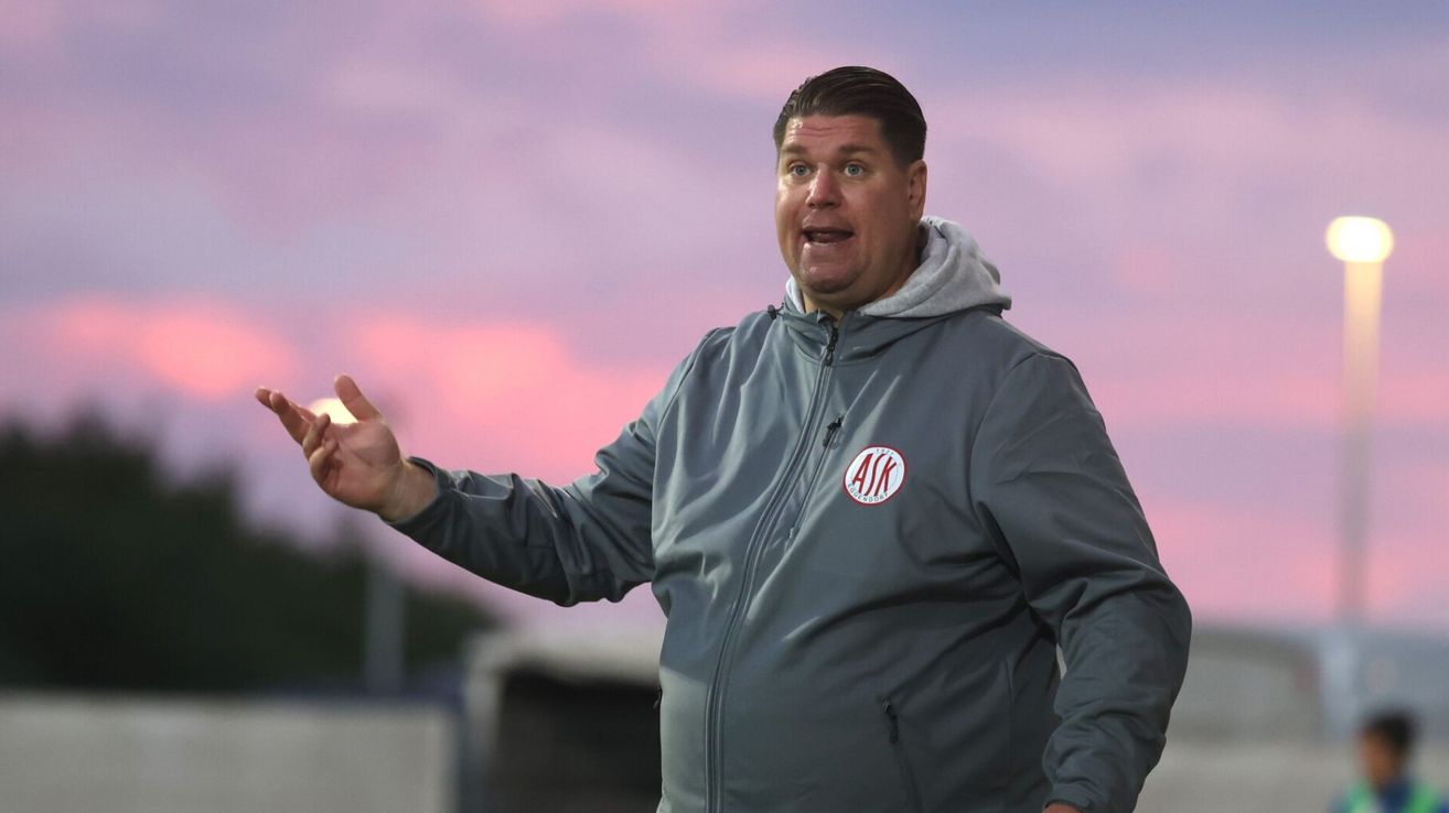 Ein Mann in einer grauen Jacke mit Logo steht auf einem Feld in der Dämmerung. Er gestikuliert mit der Hand und scheint zu sprechen.