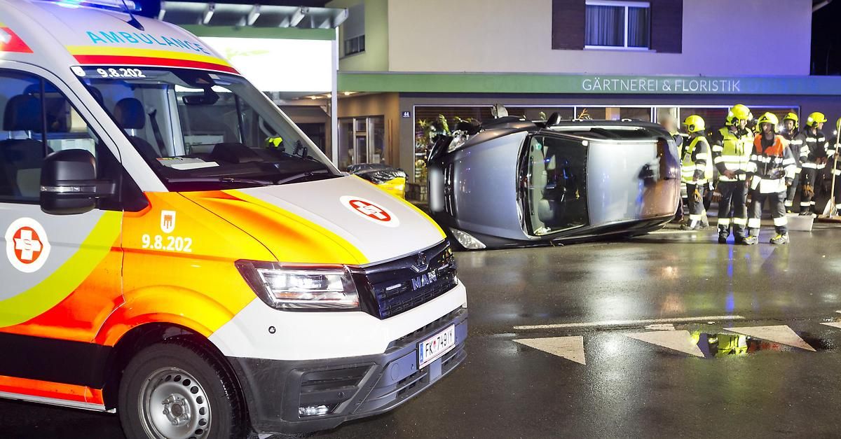 Ein Krankenwagen mit der Nummer 8.202 ist auf der Straße geparkt. Dahinter hat sich ein Auto umgedreht. Es ist Nacht, und das Gebäude hat ein Schild, das "GARTNEREI & F" besagt.