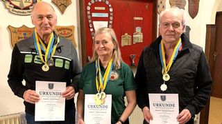 Drei Personen stehen nebeneinander, halten Urkunden in der Hand und tragen Medaillen. Sie stehen vor einer roten Wand mit verschiedenen Medaillen und Plaketten zur Schau.