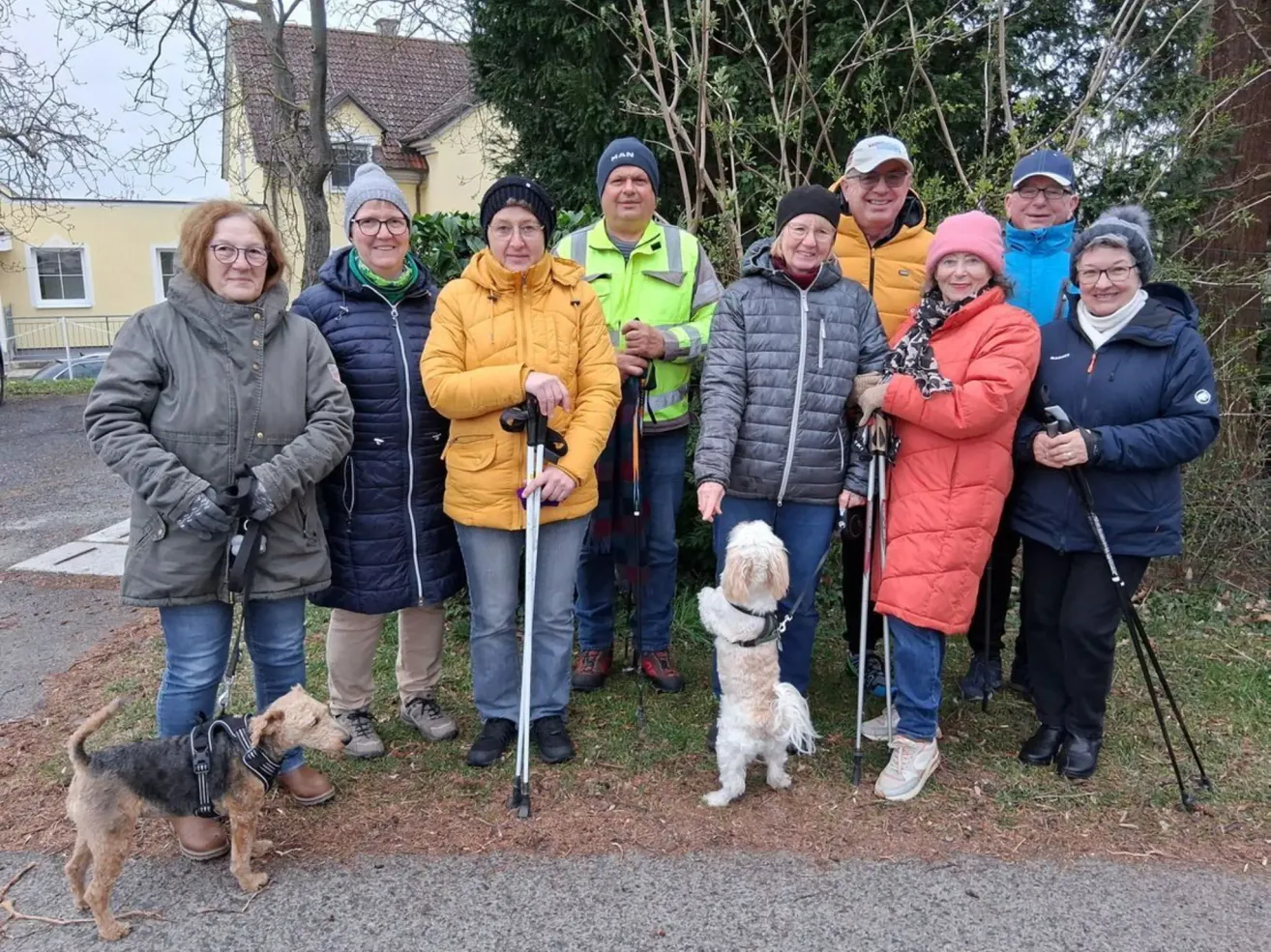 Eine Gruppe von Menschen, einige in Winterkleidung, steht in einem Park mit Bäumen und einem Haus im Hintergrund. Sie halten Wanderstöcke und haben Hunde an der Leine.