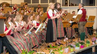 Ein Orchester spielt auf der Bühne, mit Musikern, die verschiedene Instrumente spielen. Eine Frau steht vorne und spielt Klarinette. Das Publikum sitzt dahinter, in traditionellen Kleidungsstücken gekleidet.
