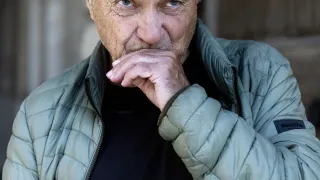 Ein grauhaariger Mann mit nachdenklichem Ausdruck. Er trägt eine grüne Jacke und ein schwarzes T-Shirt, und seine Hand ruht auf seinem Kinn.