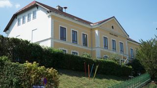 Ein gelbes Gebäude mit mehreren Fenstern und einem braunen Dach befindet sich auf einem Hang. Es trägt den Namen "VOLKS-SCHULE" oben. Vor dem Gebäude stehen ein Busch und ein Zaun. Ein Straßenschild ist auf der linken Seite des Bildes platziert.