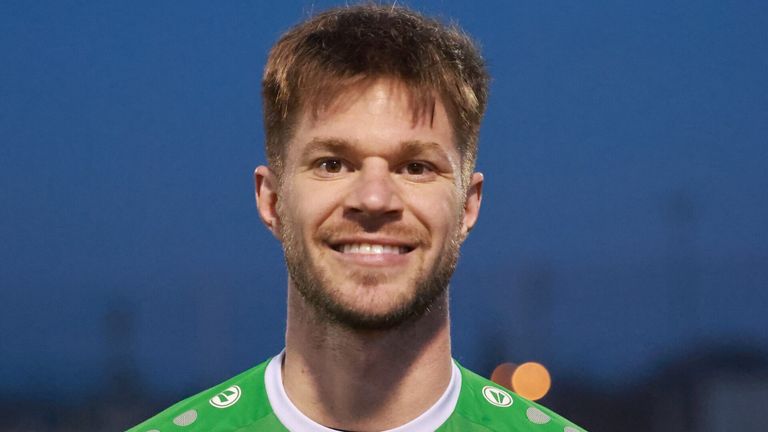 A man in a green jersey smiles at the camera with a bright blue sky in the background.