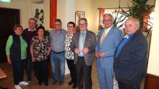 Sechs Menschen in formeller Kleidung stehen in einem Raum, möglicherweise für ein Gruppenfoto. Es gibt Topfpflanzen im Raum.