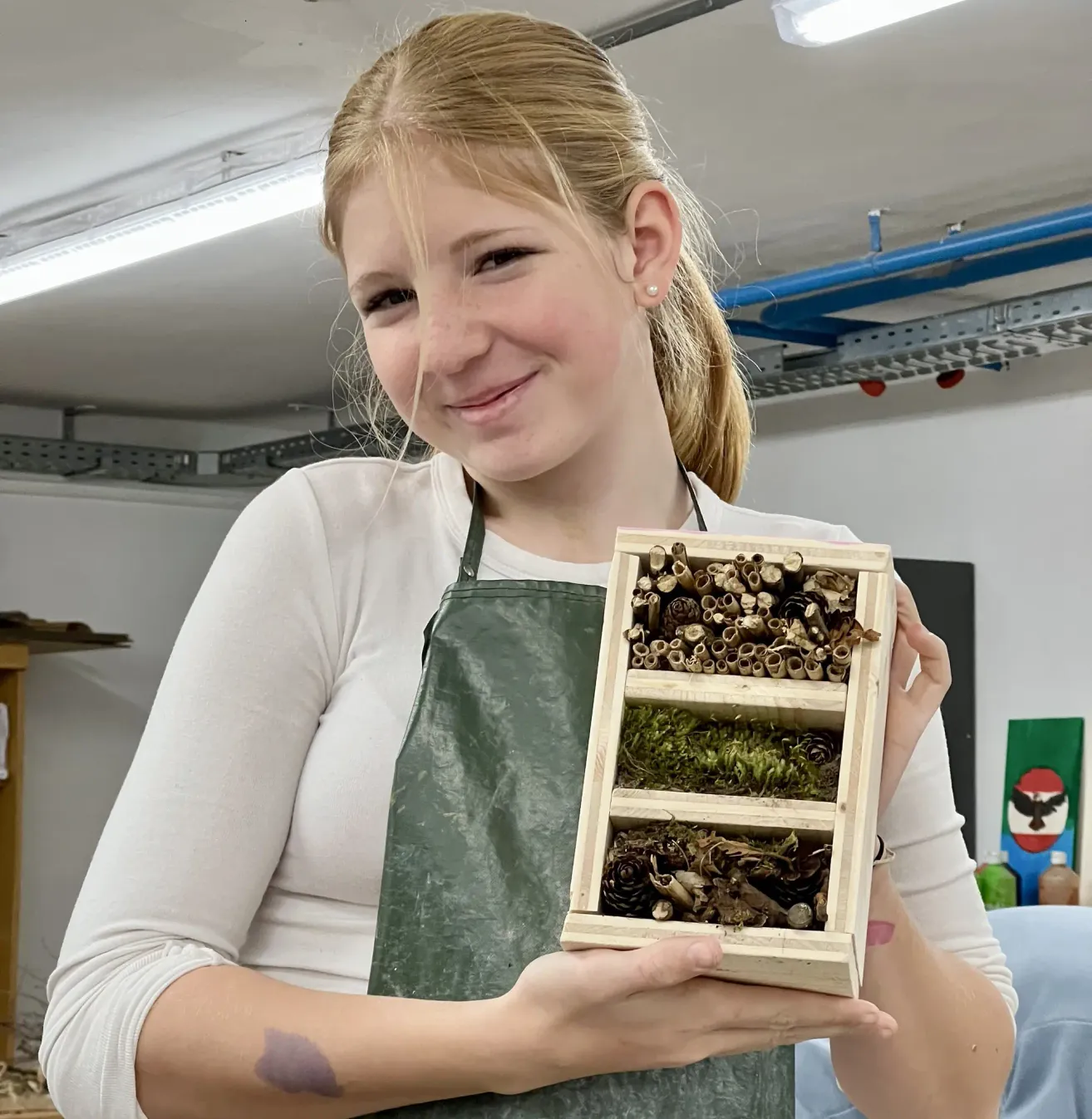Eine Frau in einer Schürze lächelt, während sie eine Holzkiste mit Tannenzapfen und Moos in der Hand hält. Sie befindet sich in einem Raum mit weißen Wänden und blauen Rohrleitungen.