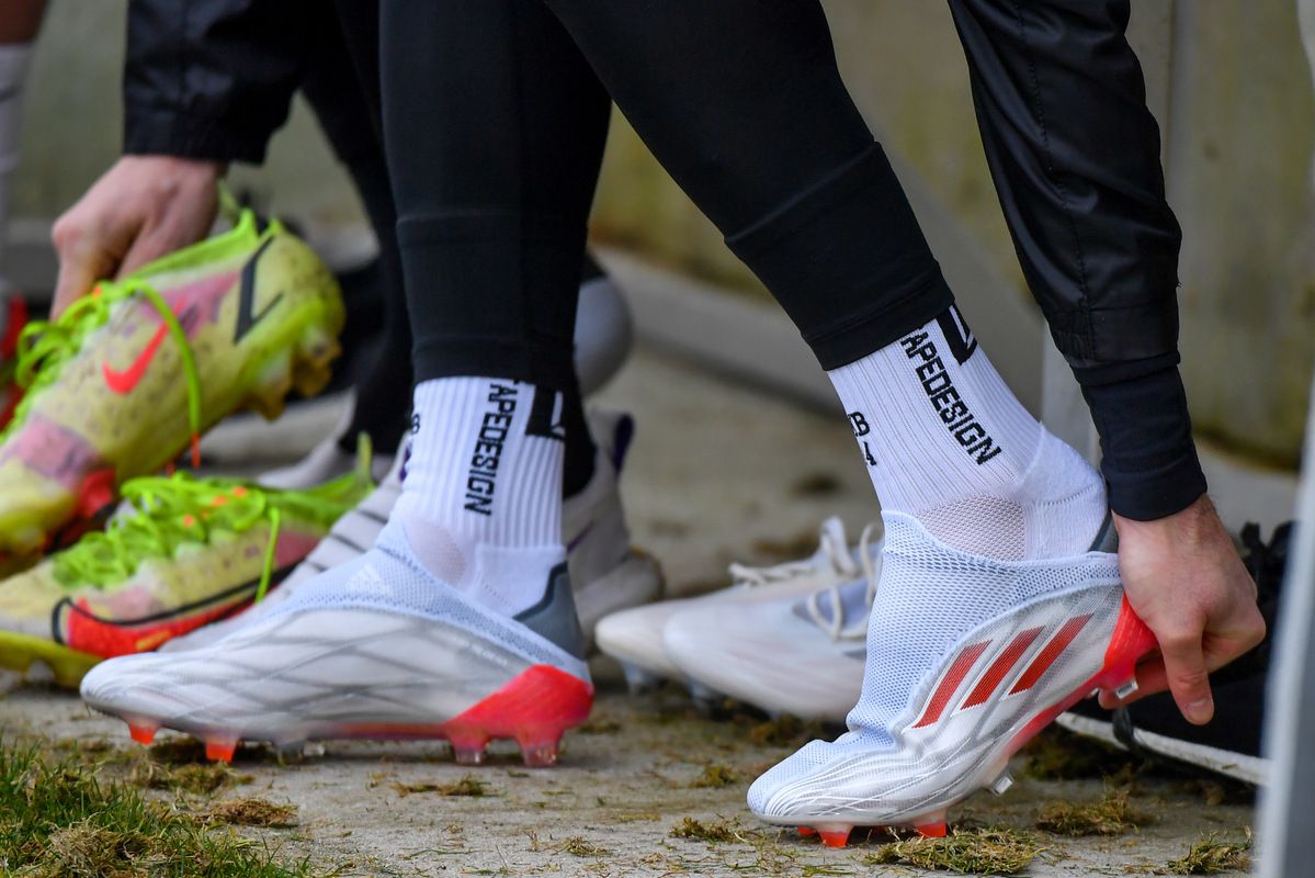 Eine Gruppe von Fußballspielern steht auf dem Feld und trägt weiße Schuhe mit roten Akzenten. Sie tragen schwarze Socken mit weißen Schriftzügen.