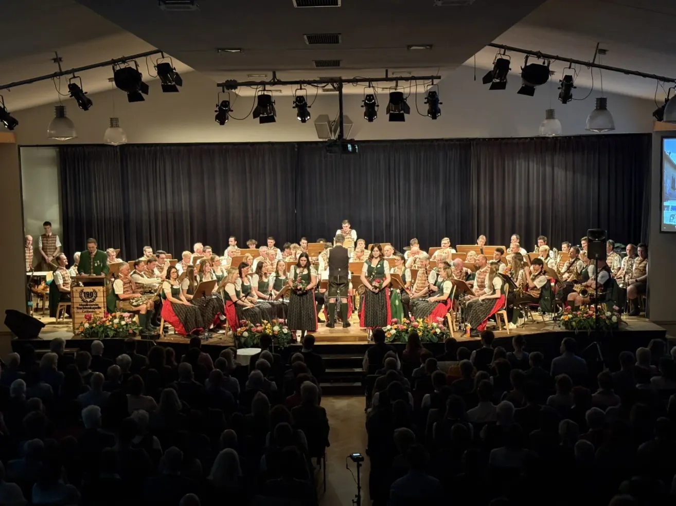 Ein großes Orchester tritt mit vielen Musikern in traditionellen grünen und roten Kleidern auf der Bühne auf, unter Scheinwerfern und vor einem sitzenden Publikum.