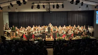 Ein großes Orchester tritt mit vielen Musikern in traditionellen grünen und roten Kleidern auf der Bühne auf, unter Scheinwerfern und vor einem sitzenden Publikum.