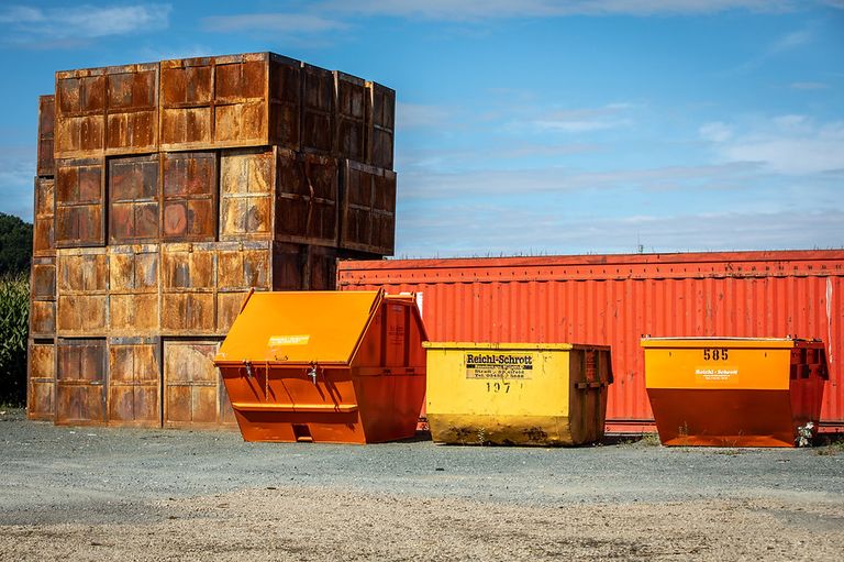 Stacks of rusty containers, including one labeled 'Reichl-Schrott,' sit against a red container and a cloudy sky.