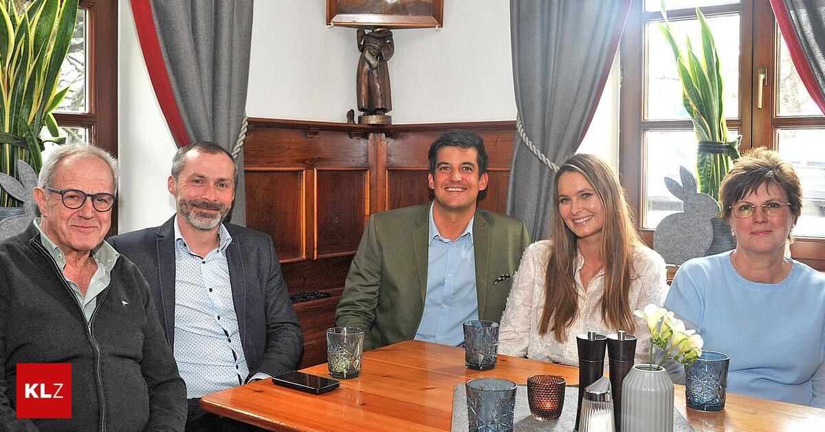 Drei Menschen sitzen lächelnd an einem Tisch. Dahinter befindet sich ein hölzerne Regal, das eine Skulptur hält. Auf dem Tisch sind ein Handy und eine Glasvase mit weißen Blumen.