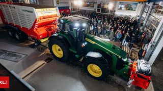 Ein grüner John Deere Traktor mit einem roten PTO wird in einem Innenraum ausgestellt, mit einem Publikum, das sich um ihn herum versammelt hat.