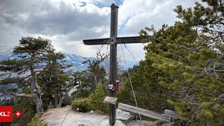 Ein holzernes Kreuz steht auf einem Berggipfel mit Blick auf ein Tal und schneebedeckte Berge. Es hat eine Tafel mit den Worten '2012' und einem Stern.