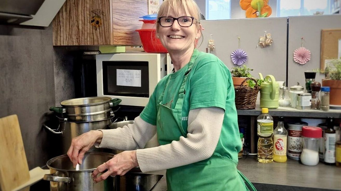 Eine Frau mit Brille und einem grünen Schürze steht in einer Küche, lächelt und hält einen Topf.