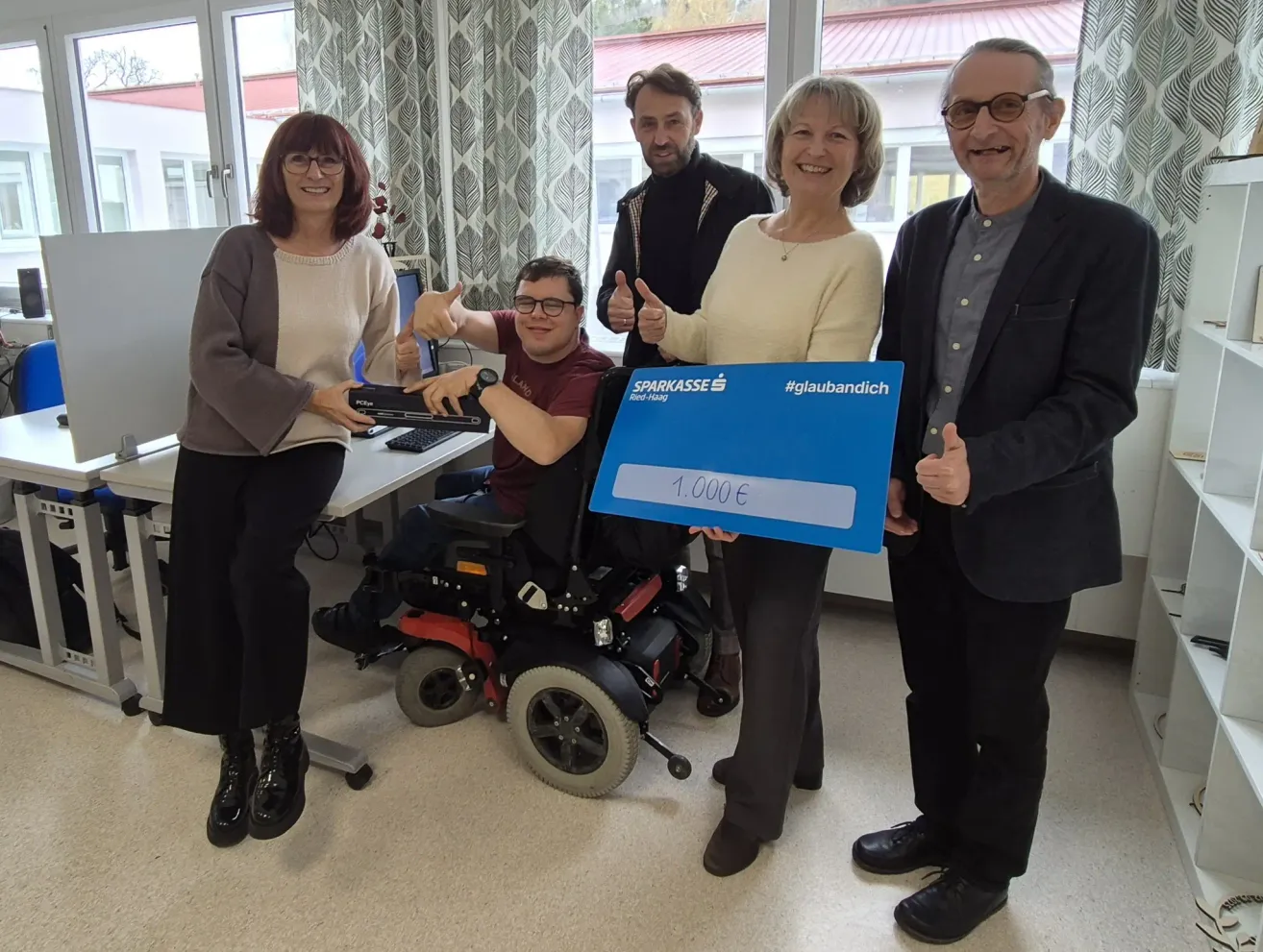 Eine Gruppe von Menschen steht in einem Raum zusammen. Ein Mann im Rollstuhl hält einen großen Scheck mit einer lächelnden Frau.