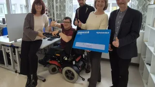 Eine Gruppe von Menschen steht in einem Raum zusammen. Ein Mann im Rollstuhl hält einen großen Scheck mit einer lächelnden Frau.