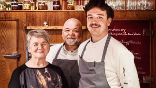 Drei Menschen in Kochschürzen stehen zusammen in einer Küche. Eine lächelnde Frau steht links, ein Mann mit Bart in der Mitte und ein lächelnder Mann rechts. Hinter ihnen sind Regale mit Gläsern und Flaschen.