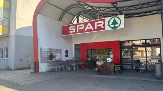 Der Eingang eines Spar-Supermarkts mit roten und weißen Wänden und einem gewölbten Dach. Das Schild darüber hat ein grünes Blatt. Eingeparkte Einkaufswagen sind draußen.