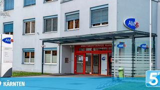 Ein weißes Gebäude mit rot umrahmten Glastüren und einem Eingangsschild mit der Aufschrift 'Arbeitsmarktservice KLAG'. Über dem Eingang befindet sich ein kreisförmiges Logo mit der Aufschrift 'AMS'.