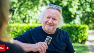 Ein Mann mit weißem Haar und Bart trägt ein blaues T-Shirt und hält ein Mikrofon. Er hat eine Sonnenbrille auf dem Kopf.
