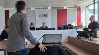 Ein Mann steht in einem Raum mit verschwommenen Gesichtern von Menschen hinter einer weißen Trennwand. Auf der Trennwand steht das Wort 'Justiz'. Ein Laptop steht auf dem Schreibtisch.
