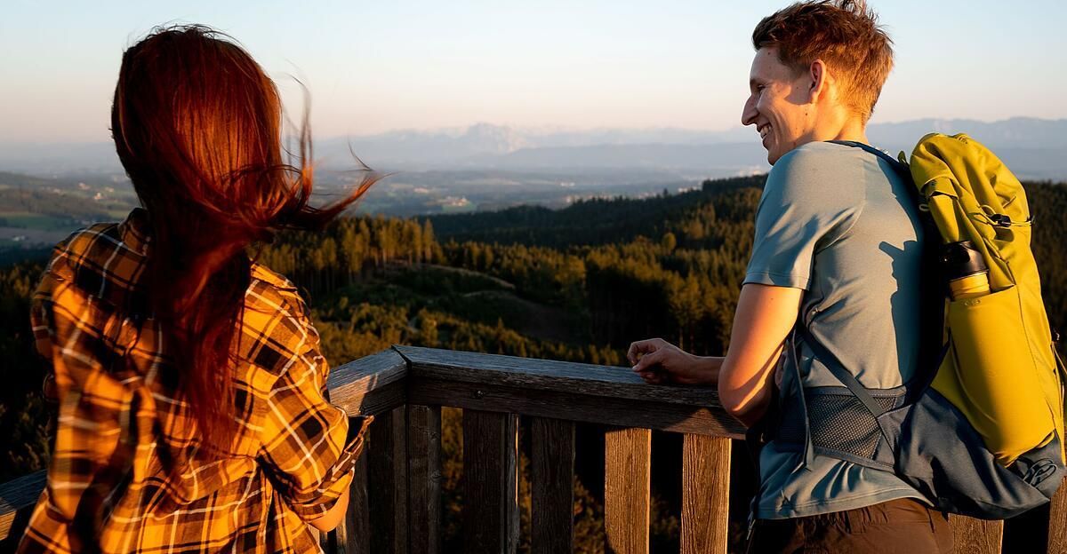 Zwei Menschen stehen auf einem Holzbalkon, lächeln und schauen auf einen Wald und Berge in der Ferne.