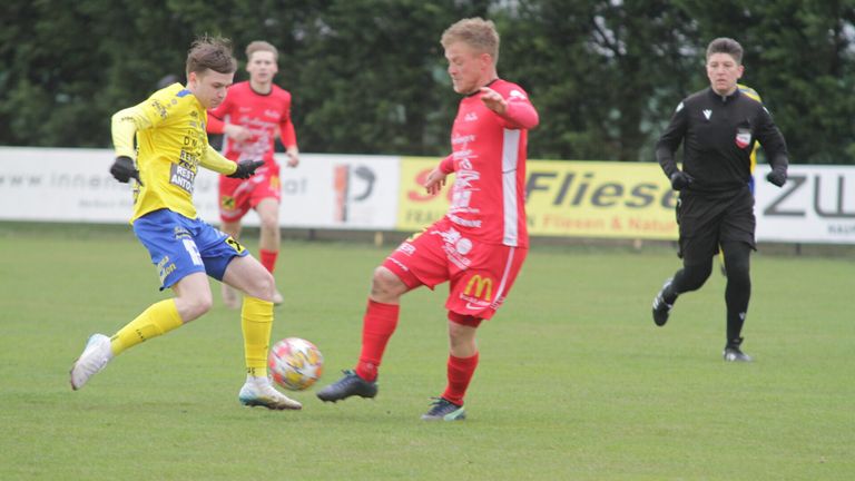 Three soccer players are on a field, two in red uniforms, one in yellow. The player in yellow is kicking the ball. Behind them is a banner with the word Flies.